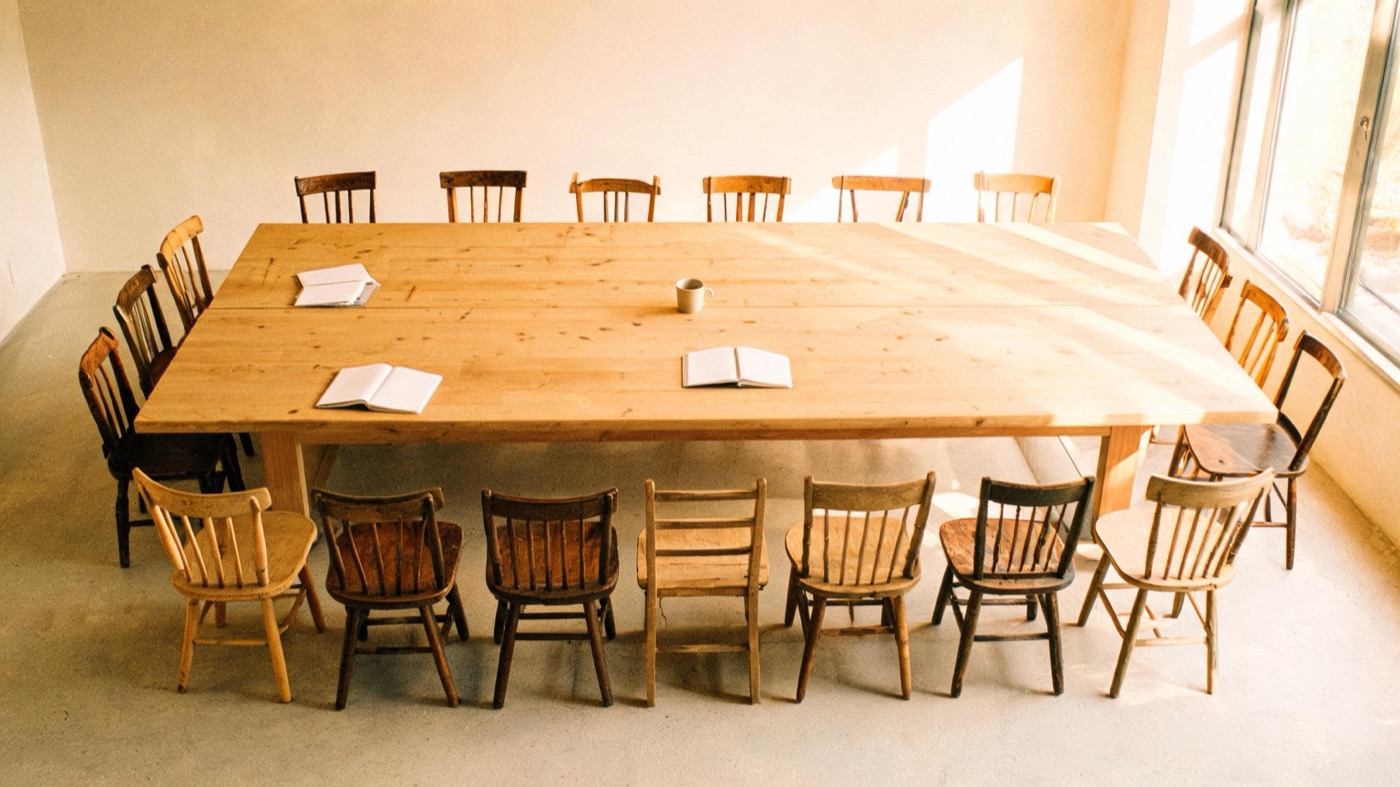 One long oak table with many mismatched wooden chairs around it, in warm morning light